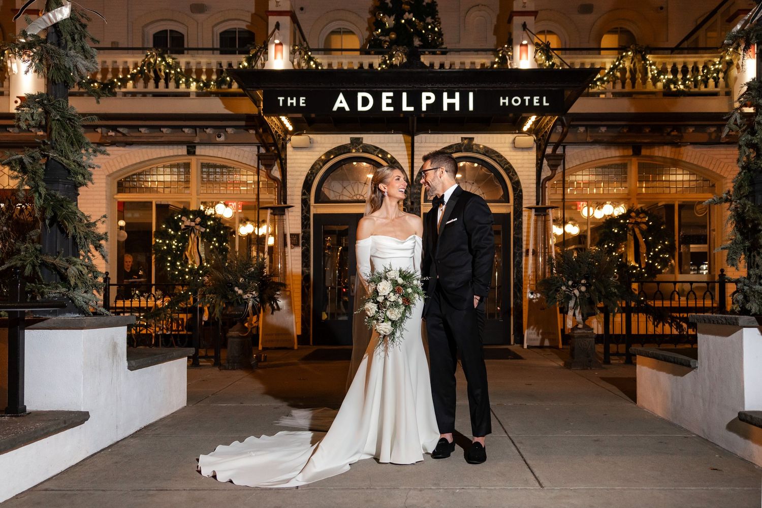 Bride and groom pose outside The Adelphi Hotel in Saratoga Springs, NY at night, surrounded by festive Christmas decorations.