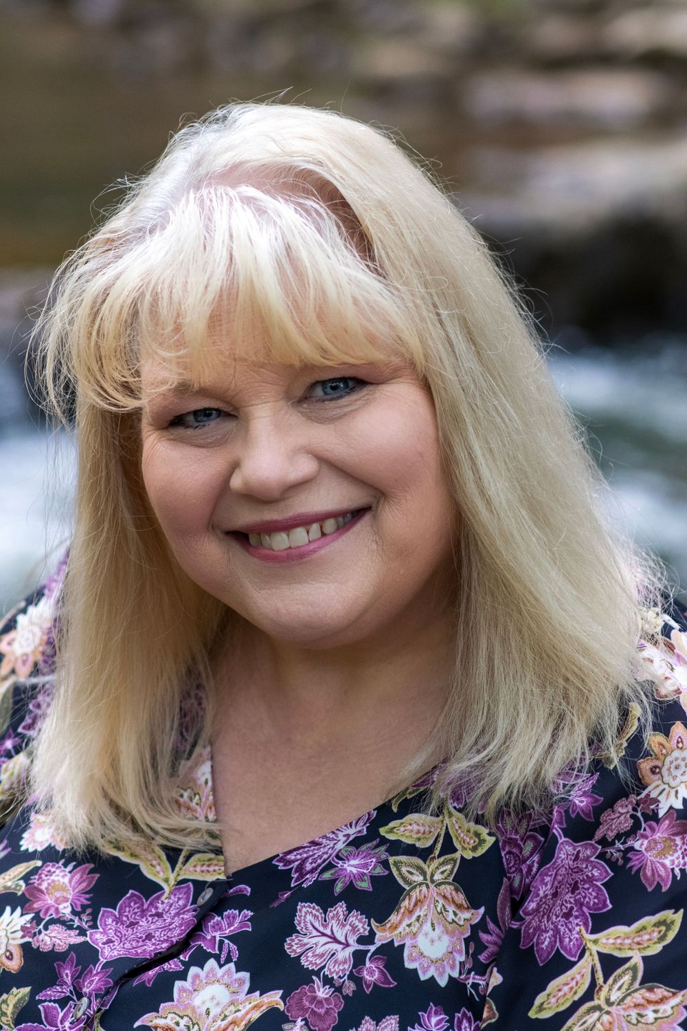 Smiling blonde woman in floral top poses outdoors near a waterfall backdrop.