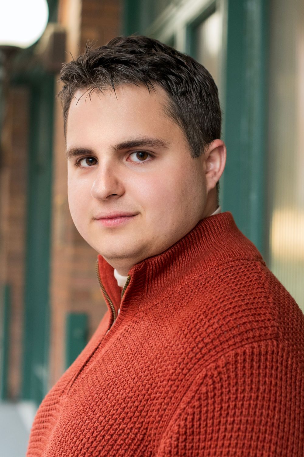 Young man in red knit sweater posing outdoors near brick building with green trim.