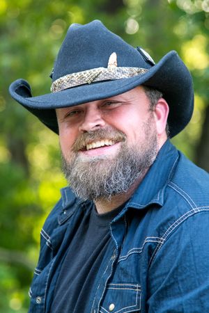 Smiling bearded man wearing a black cowboy hat and denim shirt outdoors.