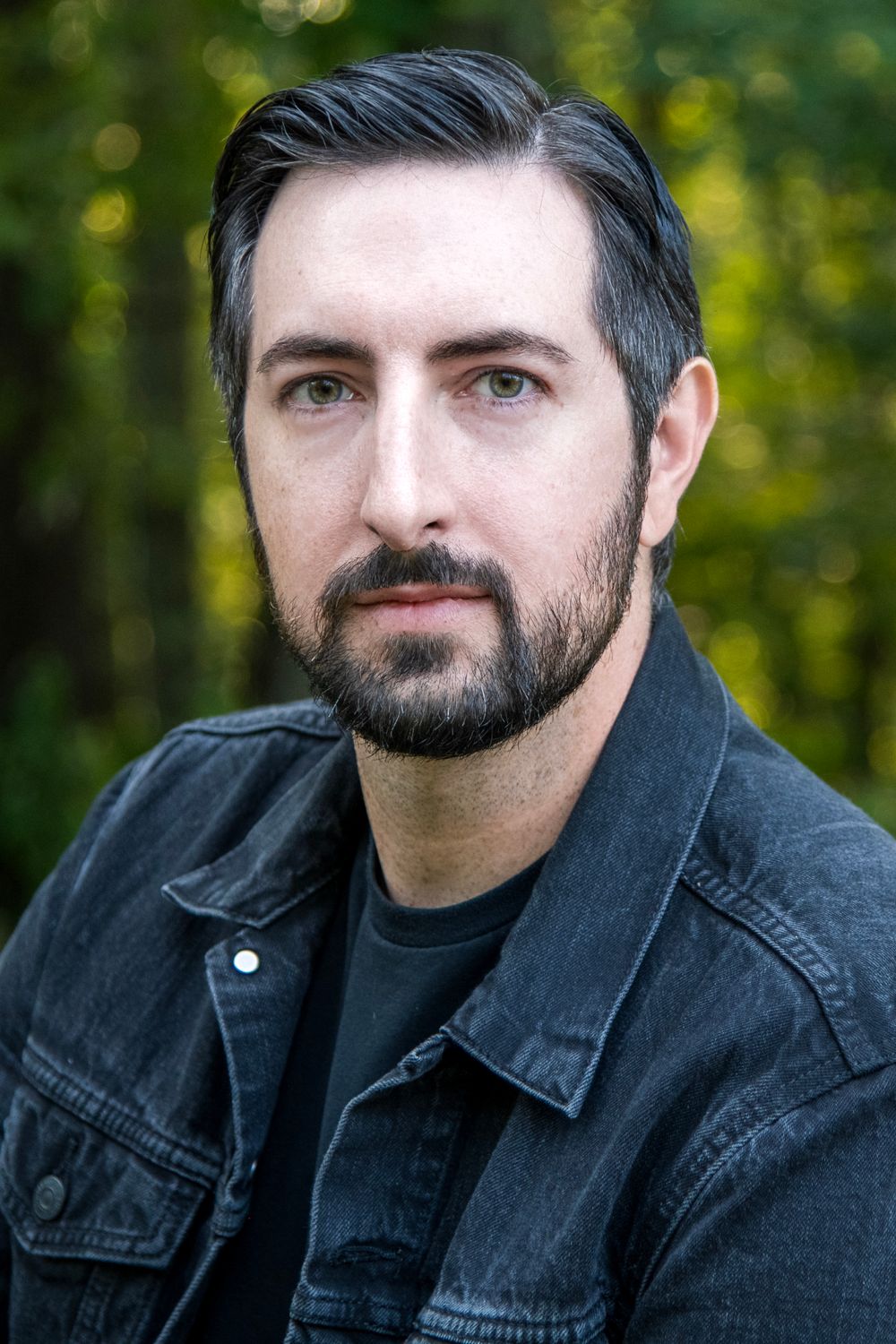Serious bearded man with dark hair wearing a black denim jacket outdoors.
