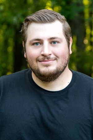 Young man with blue eyes and beard smiling outdoors, wearing black t-shirt with green trees in background.