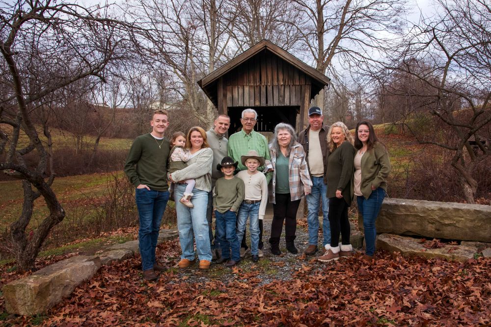 Extended multigenerational family group portrait outdoors near a rustic covered bridge in autumn.