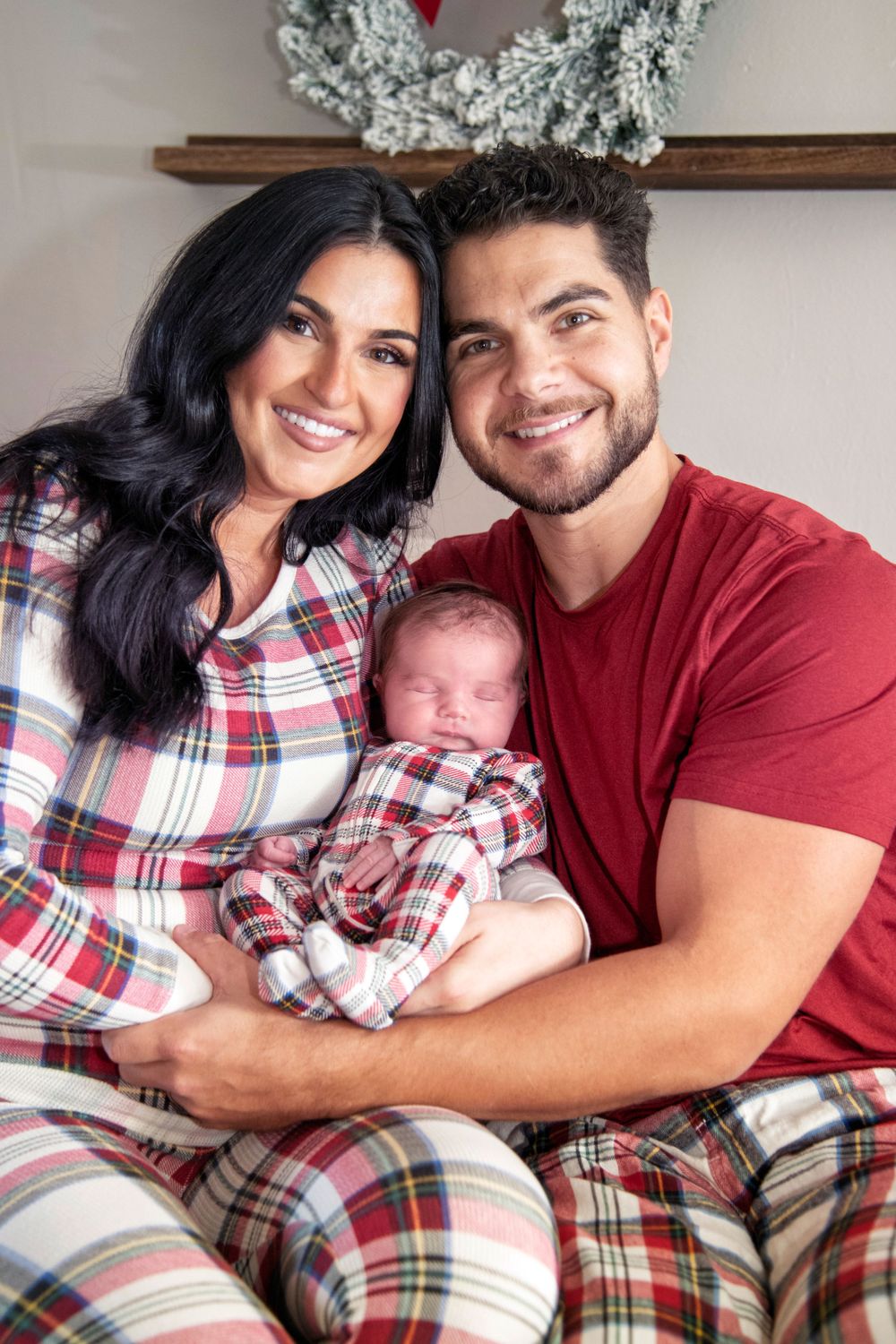 Happy young couple in plaid Christmas pajamas smiling while holding their sleeping newborn baby.