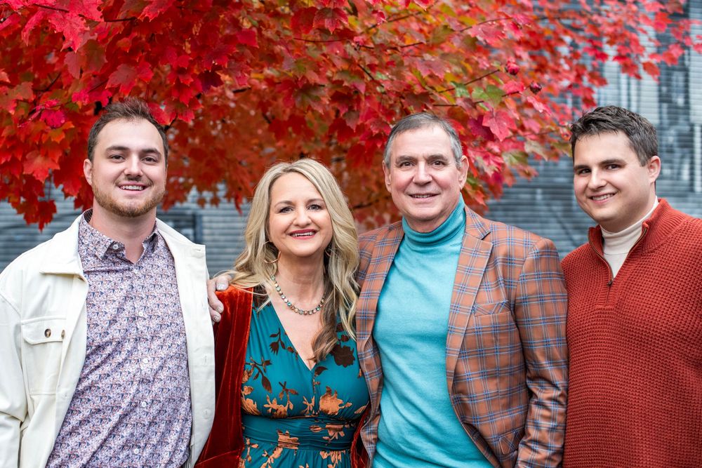 Family of four smiling together outdoors in autumn, surrounded by vibrant red fall foliage backdrop.