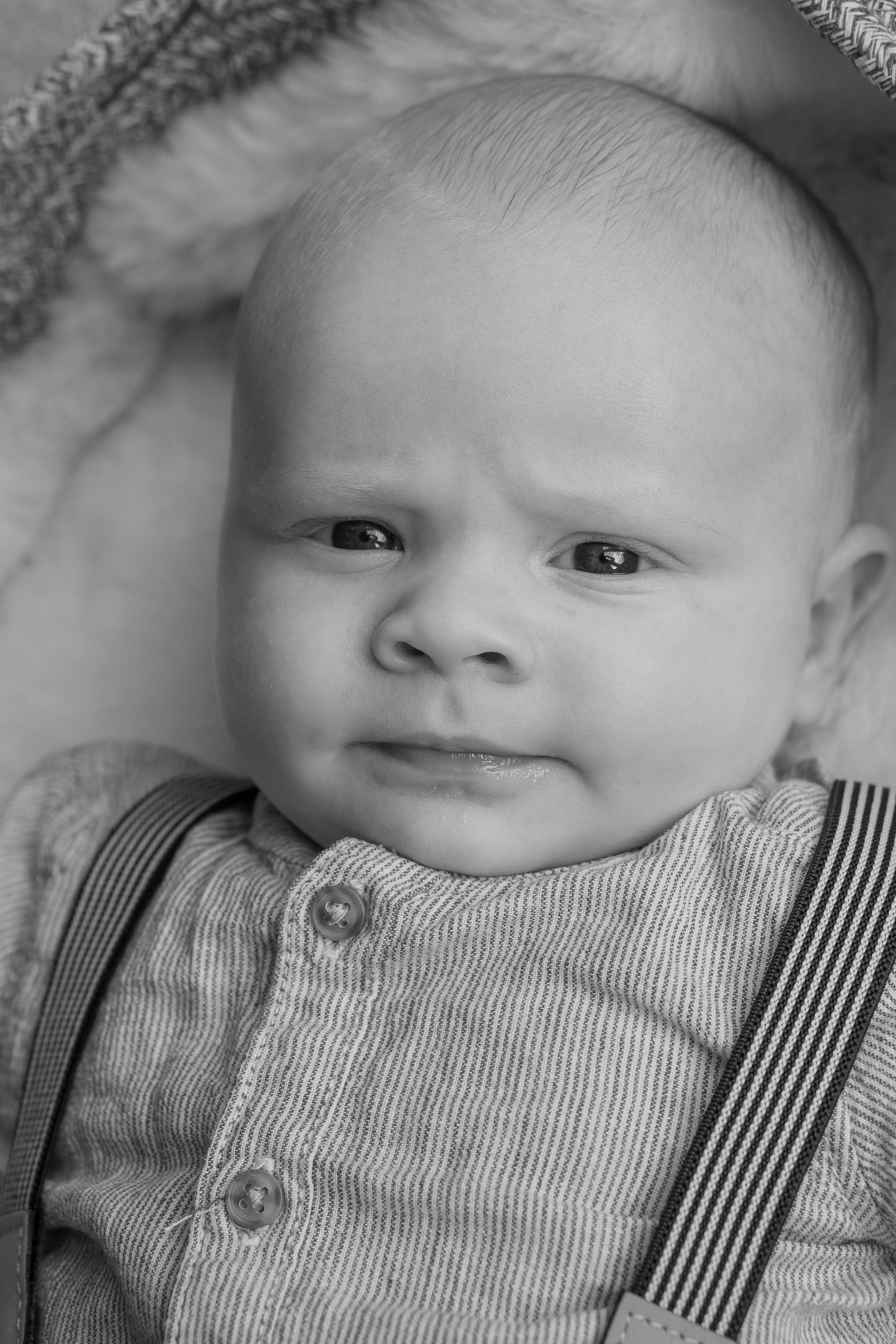 A black and white image of a newborn baby boy looking straight at the camera. He is wearing braces and a buttoned up shirt.