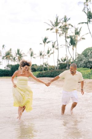 Couple holding hands and laughing while running through ocean waves on a tropical beach with palm trees.