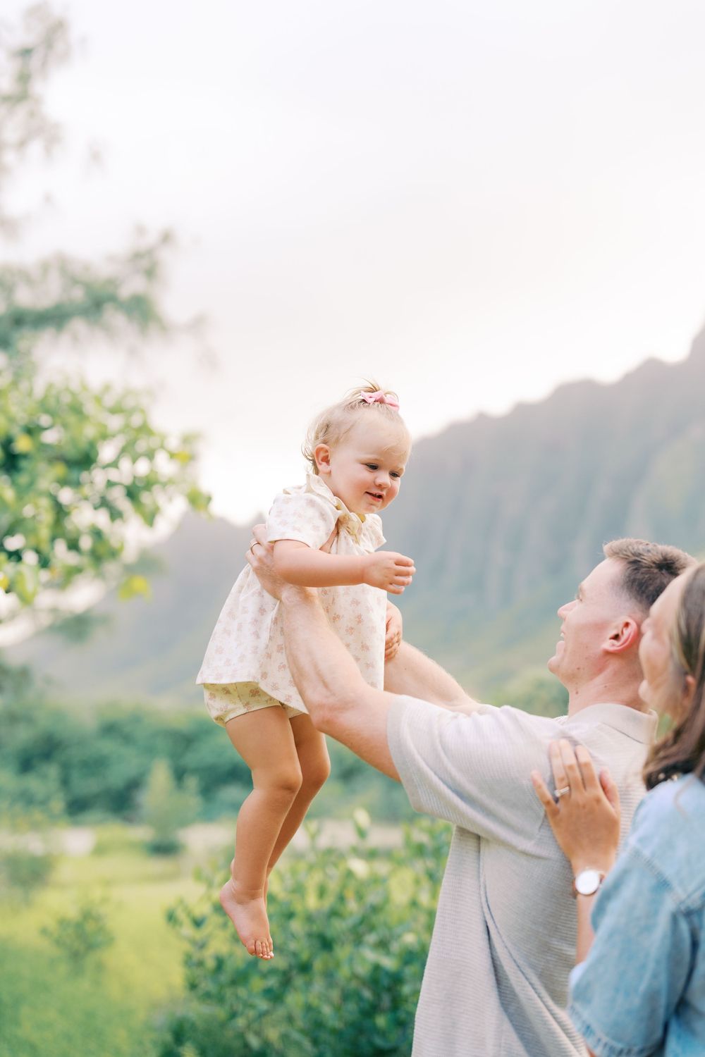 Father joyfully lifts smiling toddler girl in floral dress outdoors with lush green mountains in background.