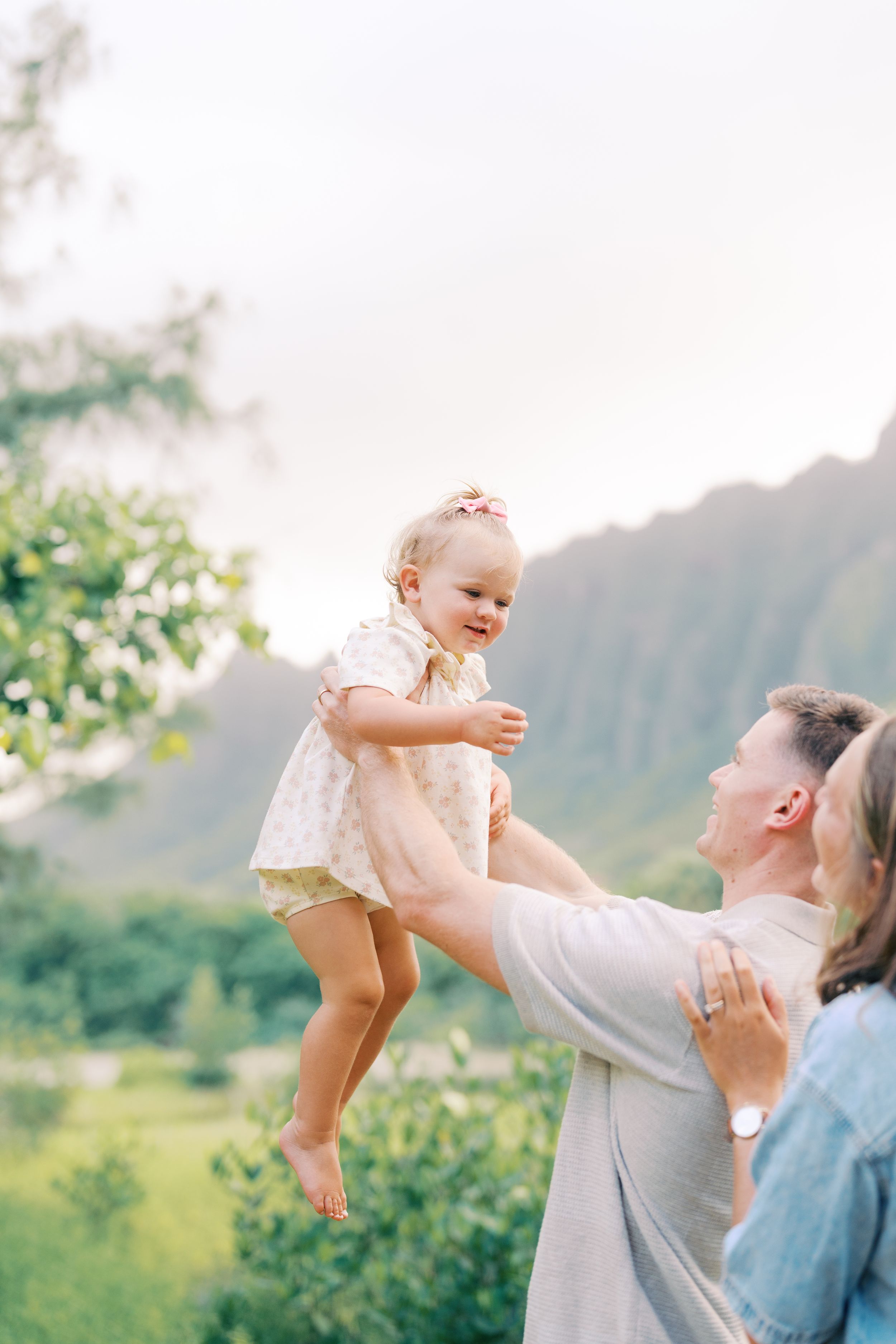 Father joyfully lifts smiling toddler girl in floral dress outdoors with lush green mountains in background.