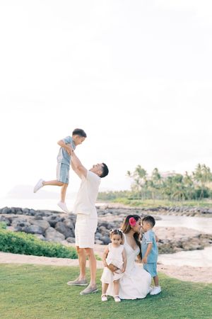 Family of five enjoys a sunny day outdoors near rocky shoreline, father tossing child in air while others smile nearby.