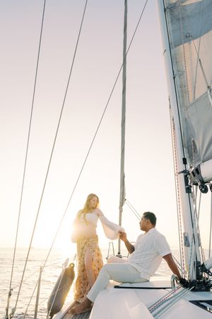 Couple holding hands on a sailboat at sunset, woman in floral dress standing while man sits on deck.