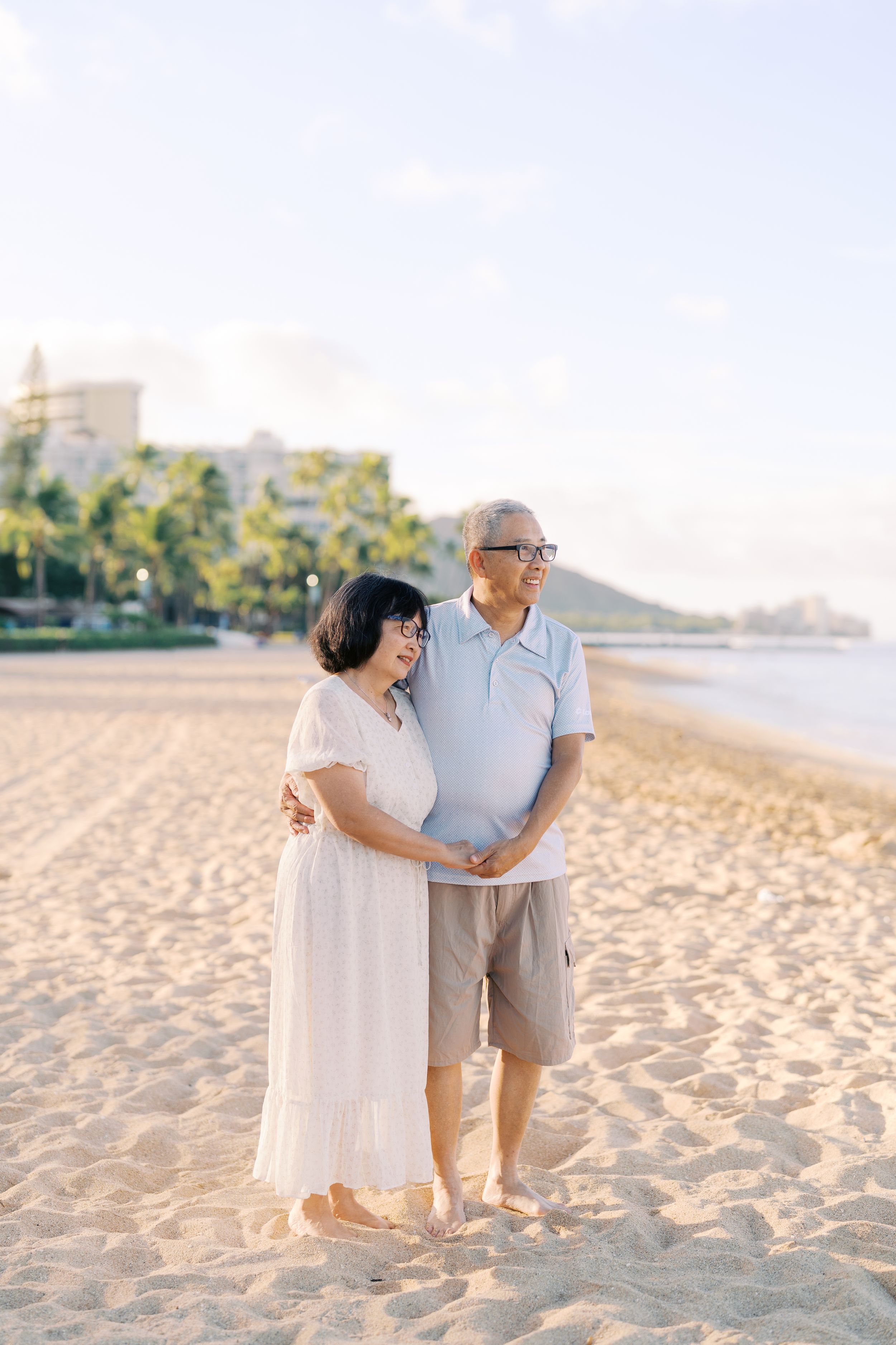 Elderly Asian couple embracing on a sunny sandy beach, with palm trees and buildings in the background.