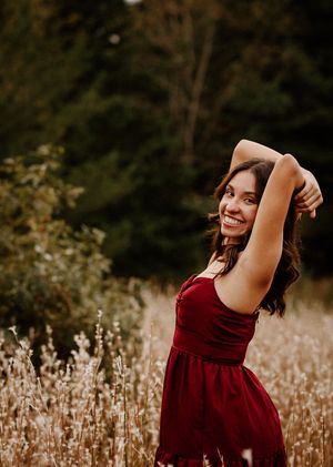 Young woman in red velvet dress smiling joyfully in a golden autumn meadow with trees in background.