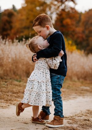 Young boy kissing his little sister on the cheek while hugging on a dirt path in autumn.