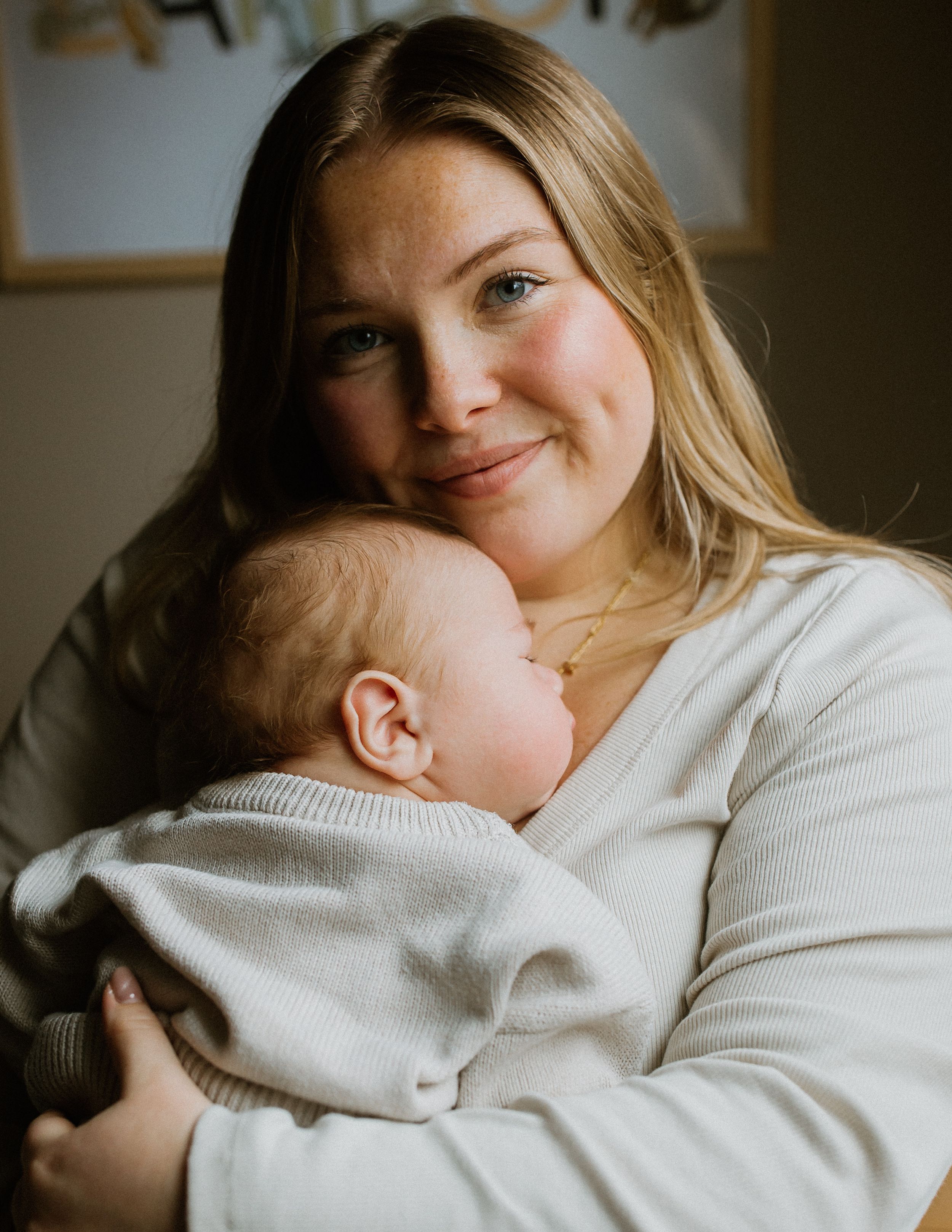 Smiling blonde mother gently holds sleeping newborn baby against her chest in a cozy nursery.