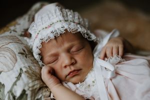 Peaceful sleeping newborn baby wearing a delicate white lace bonnet resting on a floral blanket.