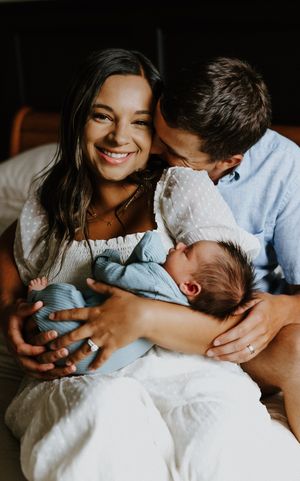 Smiling mother holding newborn in blue wrap while father kisses her cheek in warm indoor setting.