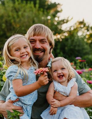Smiling father holding two blonde toddler daughters in a colorful flower garden at sunset.