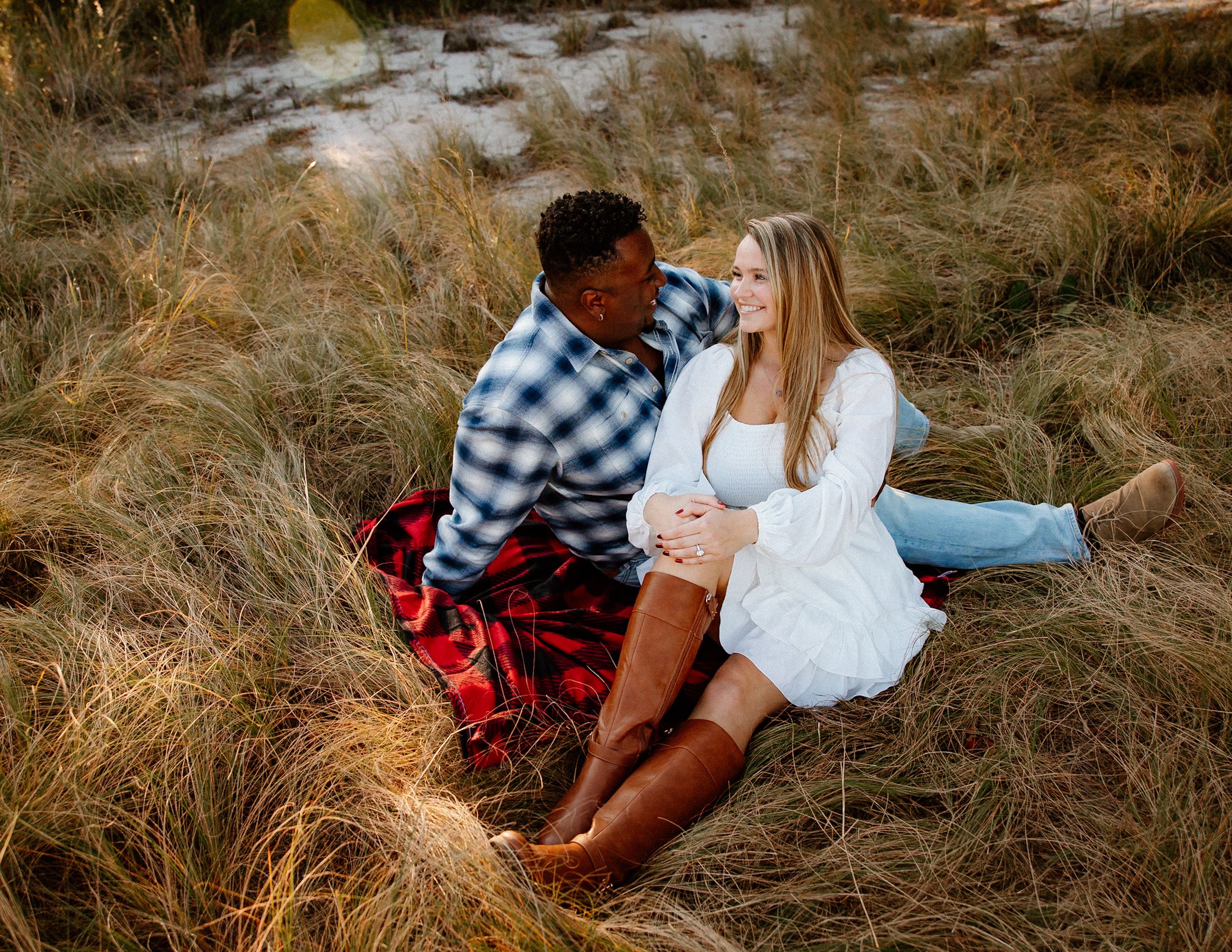 Interracial couple sitting in golden tall grass at sunset during a romantic maternity photo session.