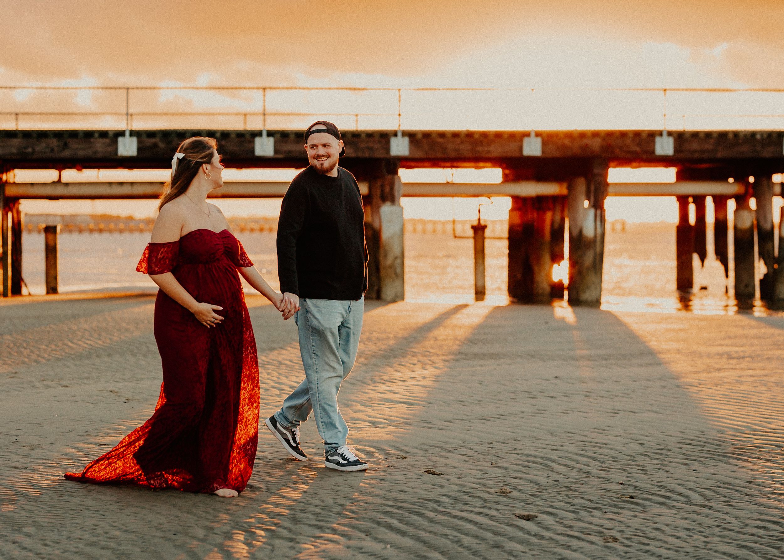 Pregnant couple holding hands walking on sandy beach at sunset near a pier, woman in red gown.