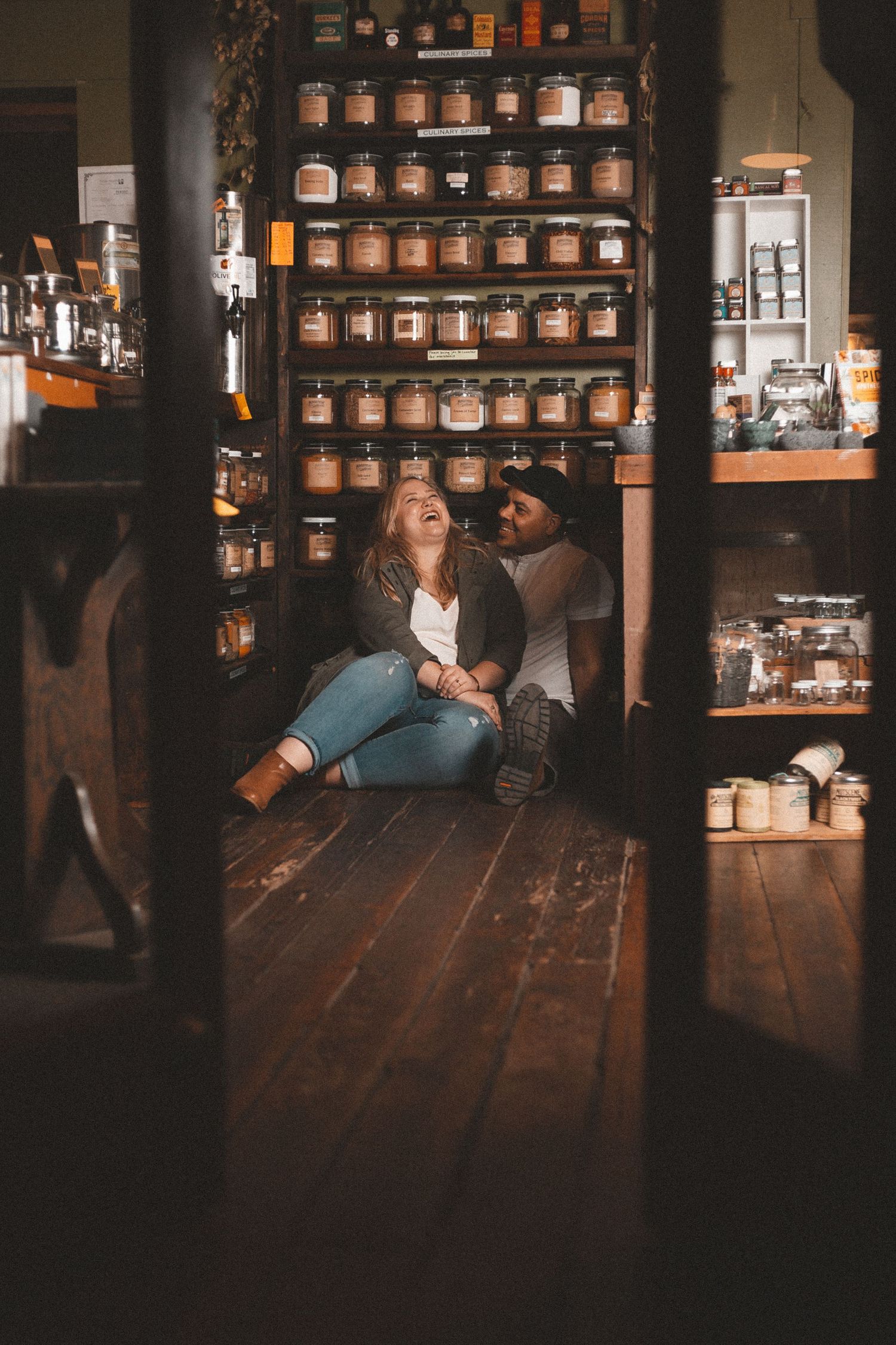 A couple sit in front of a shelf of many jars. She is sitting in his lap and laughing while he looks at her smiling.