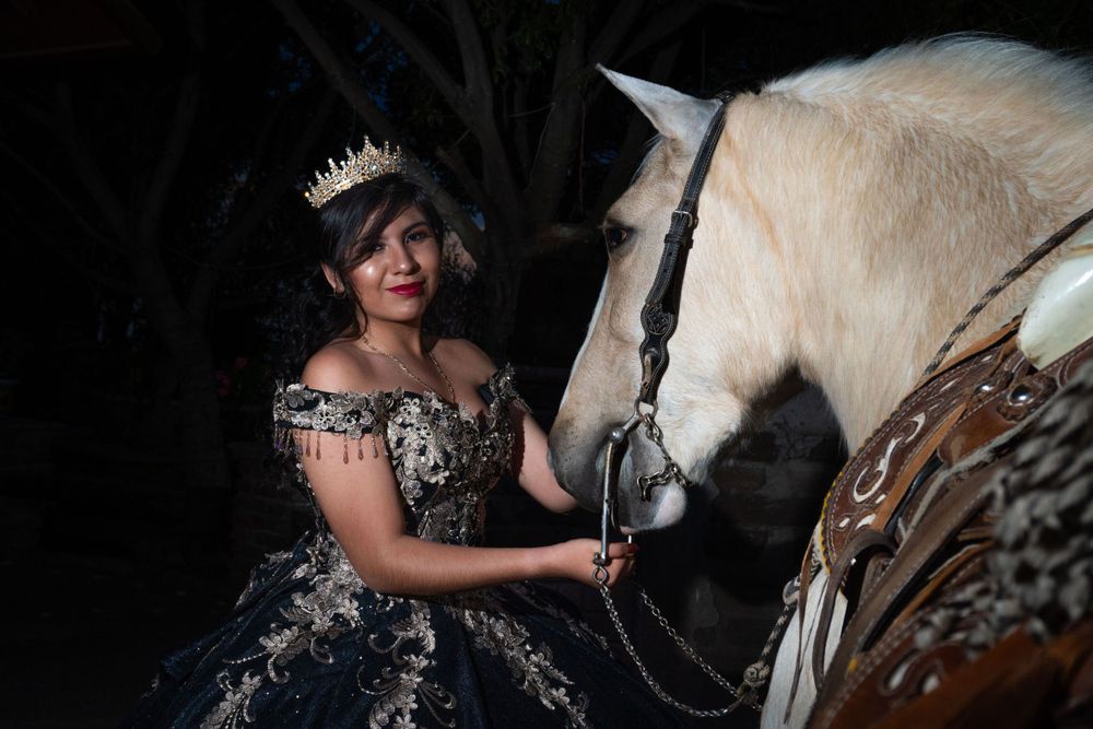 Young woman in elegant black quinceañera gown and crown posing with a white horse in dark setting.