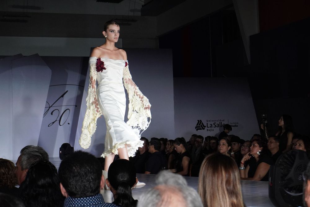 Female model in an elegant white gown with floral accents walking the runway at a fashion show.