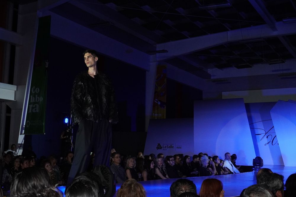 Male model walking the runway in a dark outfit under blue stage lights at a fashion show.