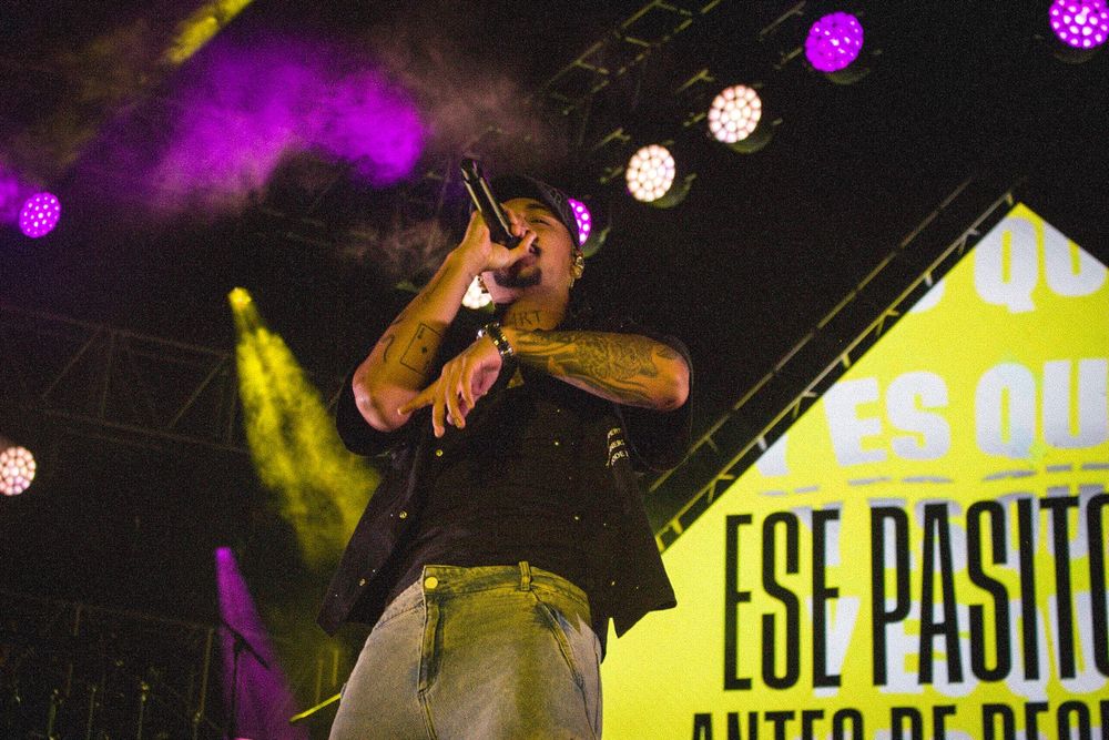 Hip hop artist performing on stage with microphone under purple and yellow stage lights at concert.