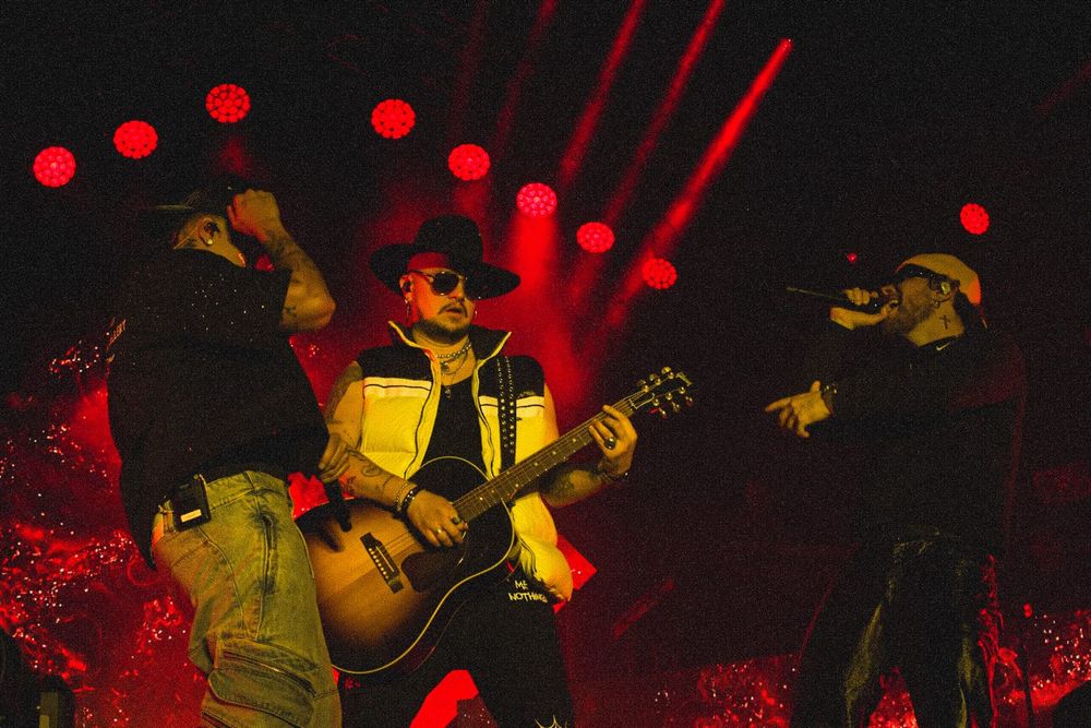 Three musicians performing on stage with guitarist in center under dramatic red concert lighting.