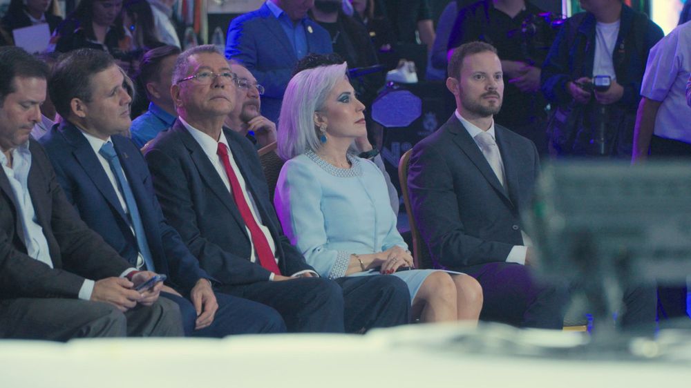 Four formally dressed professionals seated in audience at a corporate conference or business event.