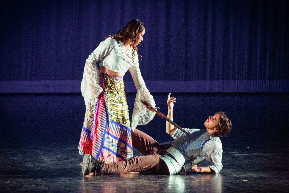 Two ballet dancers perform a dramatic scene on stage, one standing over the other who lies on the floor.