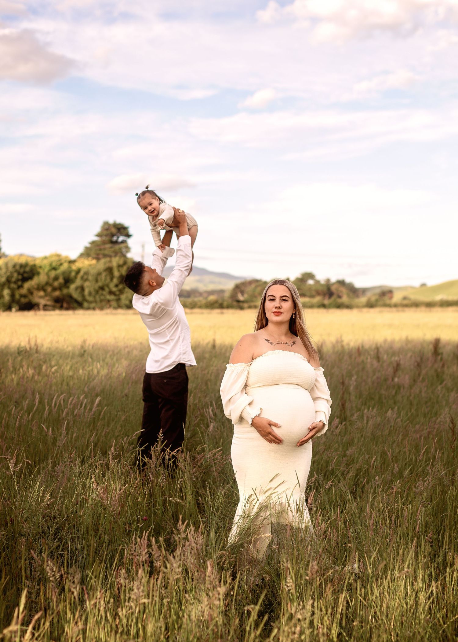 A couple poses in a golden field at sunset with a small child lifted in the air and a pregnant woman in a white dress.