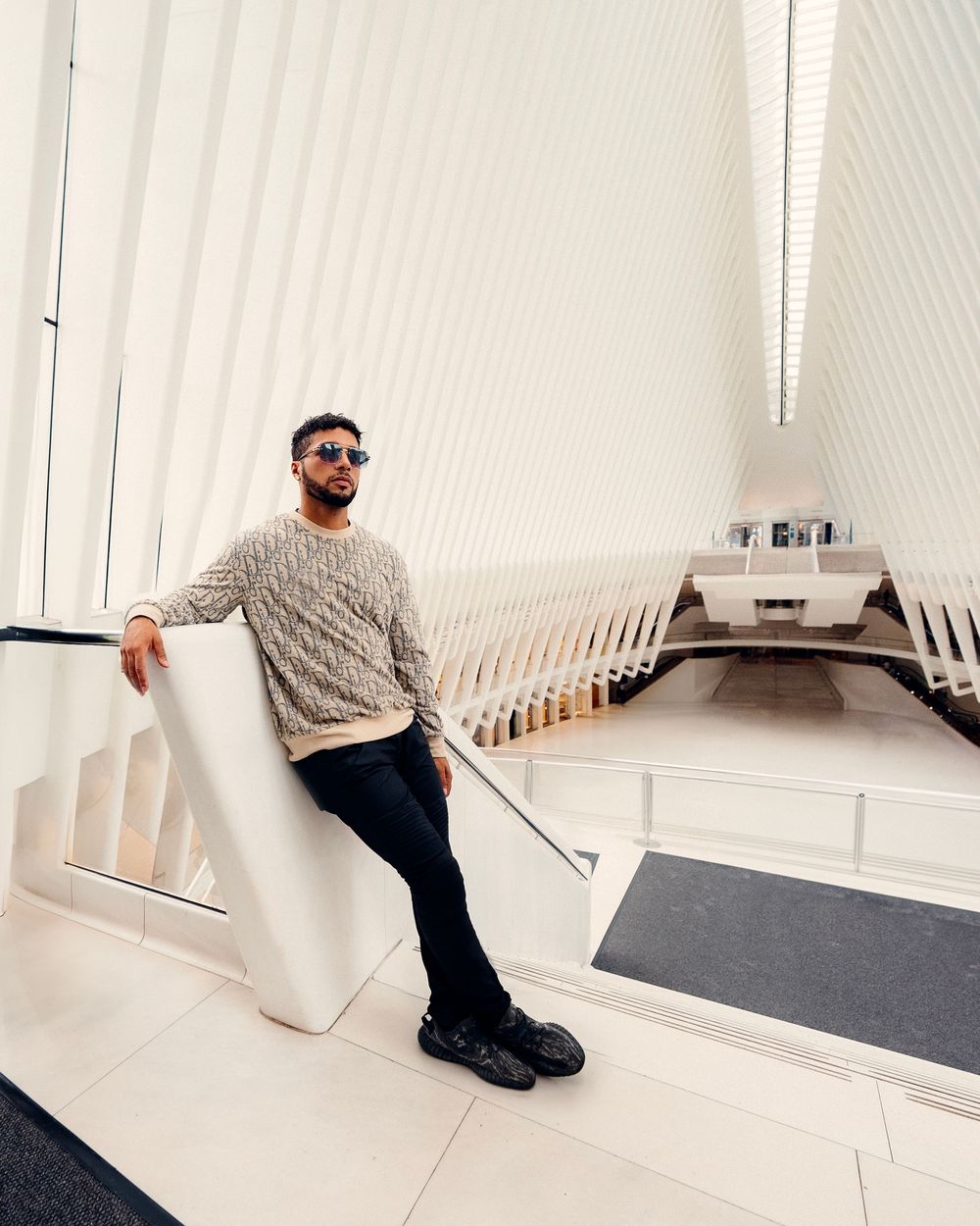 Person in white sweater and black pants reclines against white wall in minimalist architectural setting.