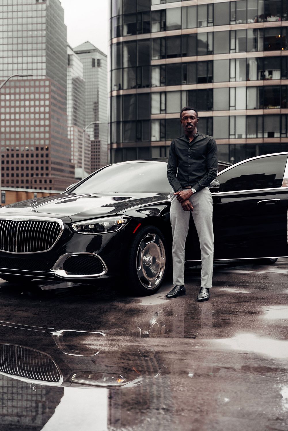 A stylish black luxury car parked in front of modern glass buildings on a rainy urban street.