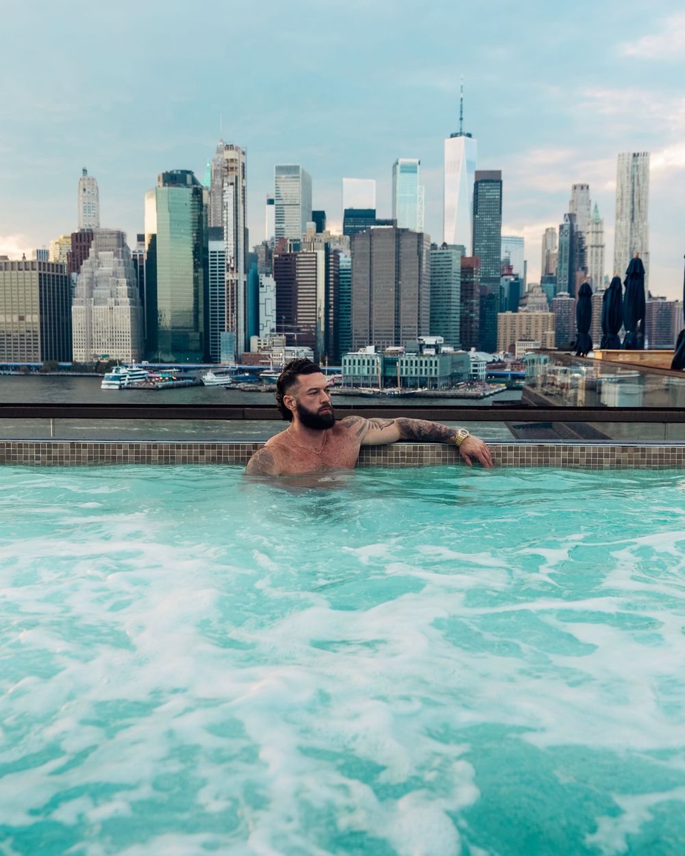 Panoramic view of a rooftop infinity pool overlooking the Manhattan skyline on a sunny day.