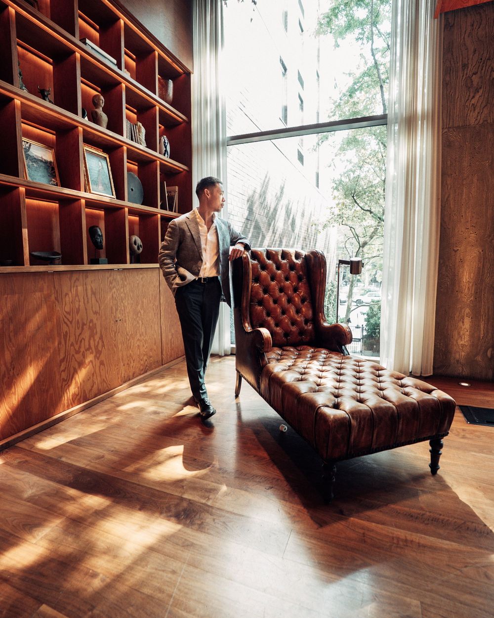 A leather chesterfield chair sits in a sunlit room with wooden floors and built-in shelving along the walls.