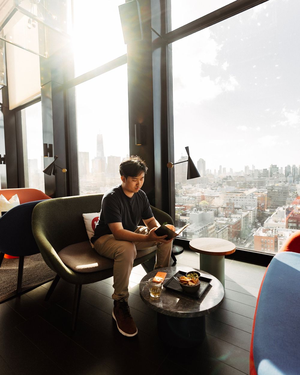 Person sitting alone at window table in modern high-rise restaurant with dramatic cityscape view.