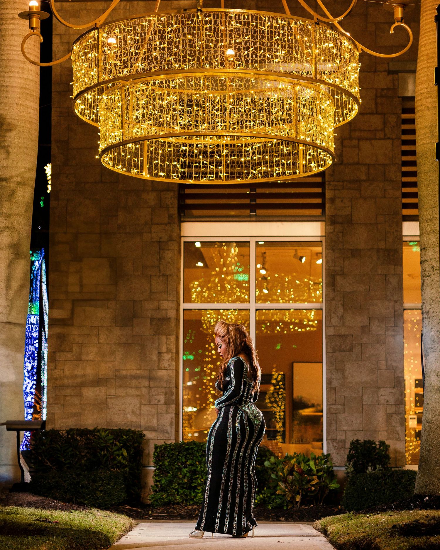 Large illuminated chandelier with cascading lights hangs above a garden entrance decorated with holiday ornaments and lighting.