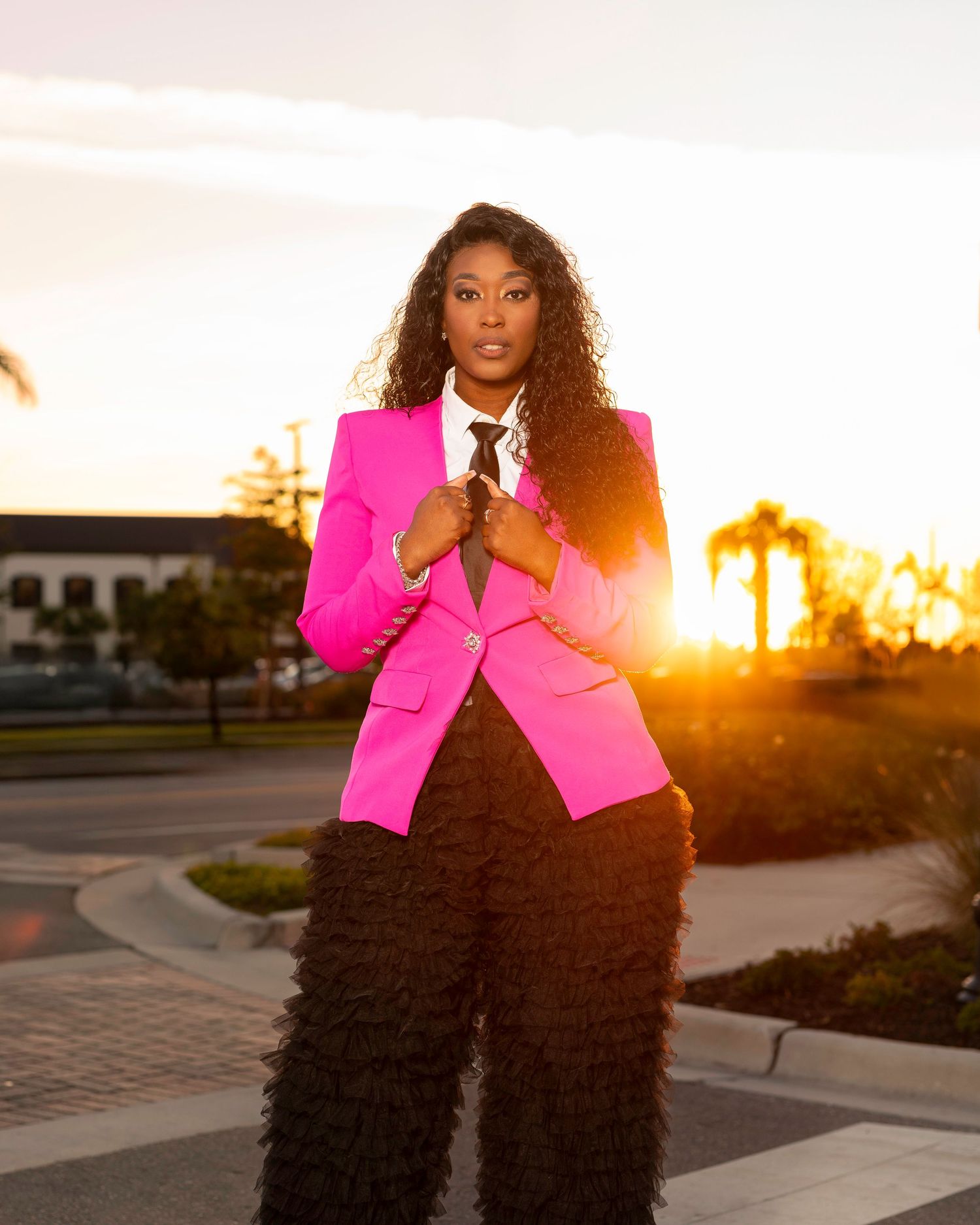 A woman in a hot pink blazer and ruffled black pants poses outdoors at sunset with palm trees in the background.