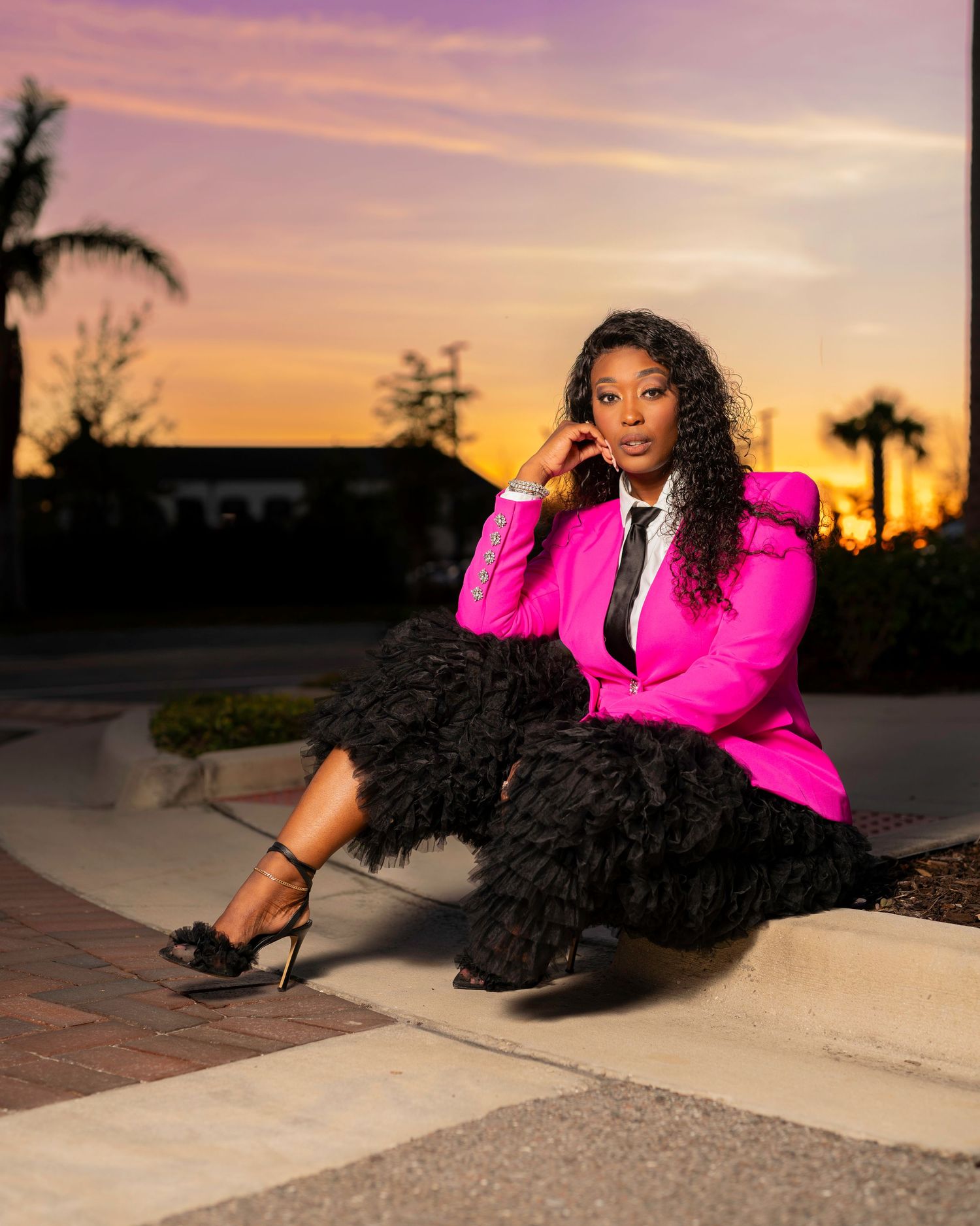 A fashion portrait of a person in a vibrant hot pink blazer and black tulle skirt sitting outdoors at sunset.