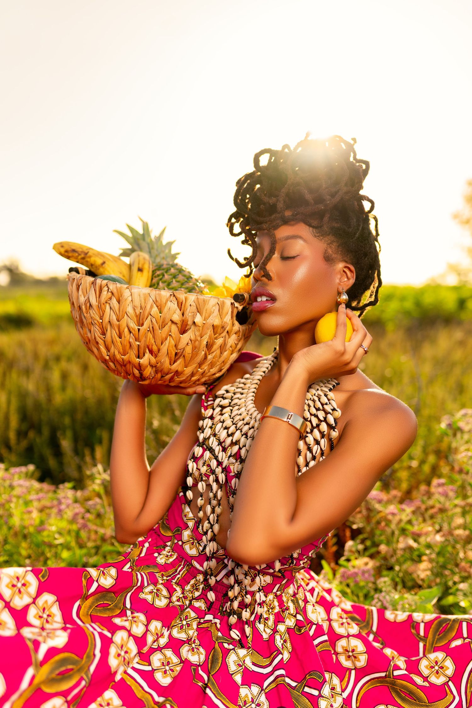 A person in a pink floral dress poses with a pineapple at sunset in a flower field.