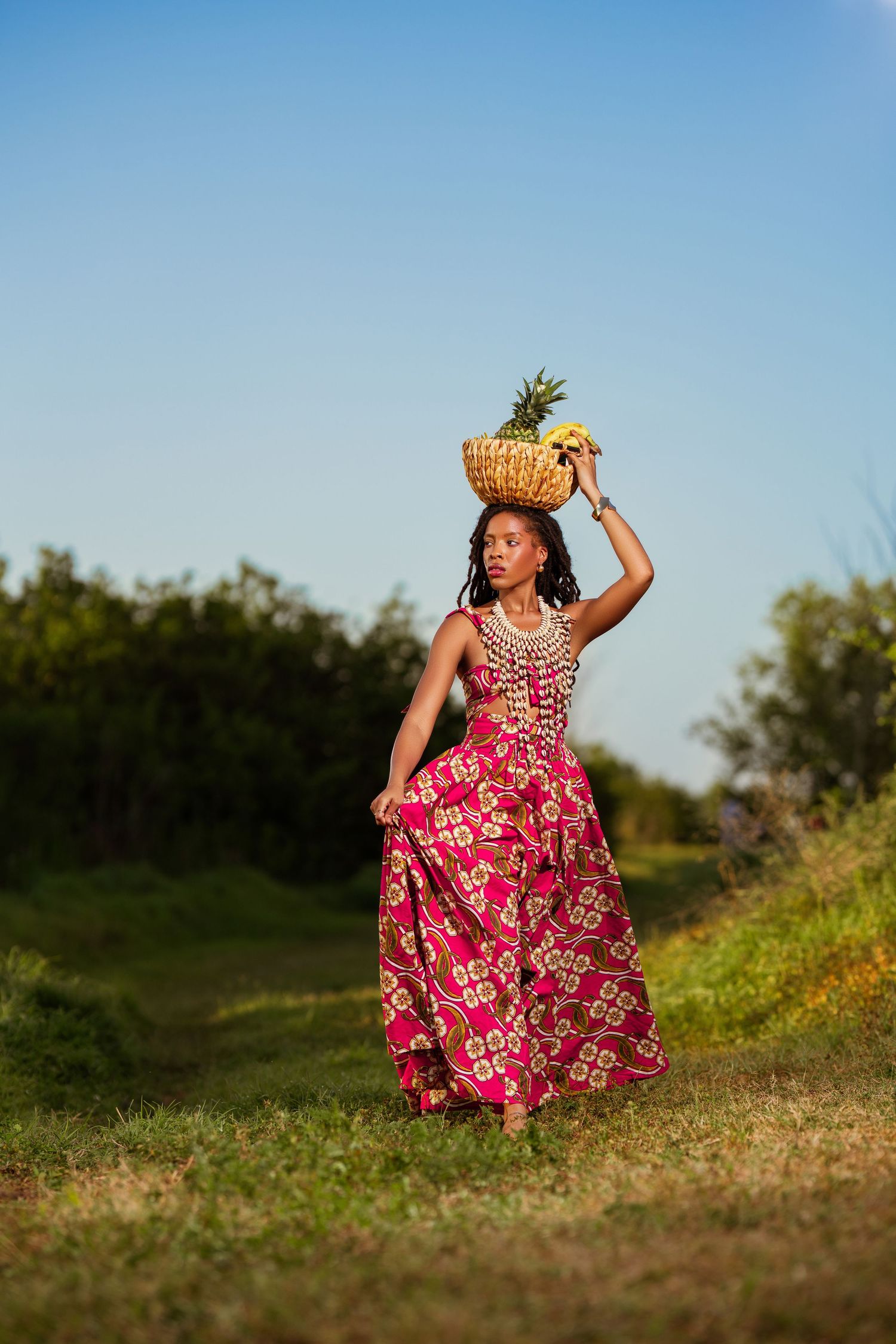 A person in a vibrant pink patterned maxi dress stands in a grassy field holding a pineapple above their head at sunset.