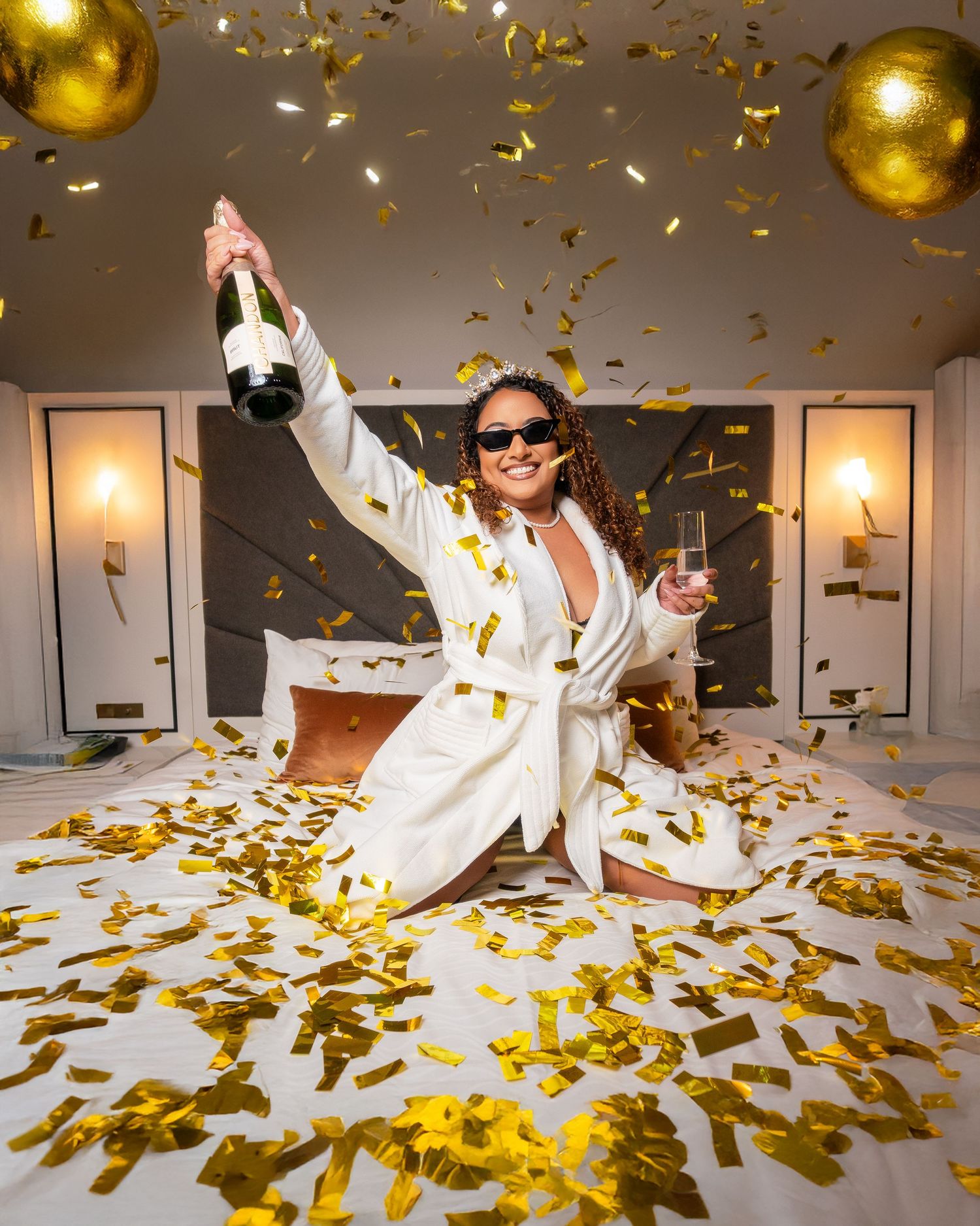 Celebratory scene with gold confetti and balloons as someone in a white robe and sunglasses holds champagne bottles.