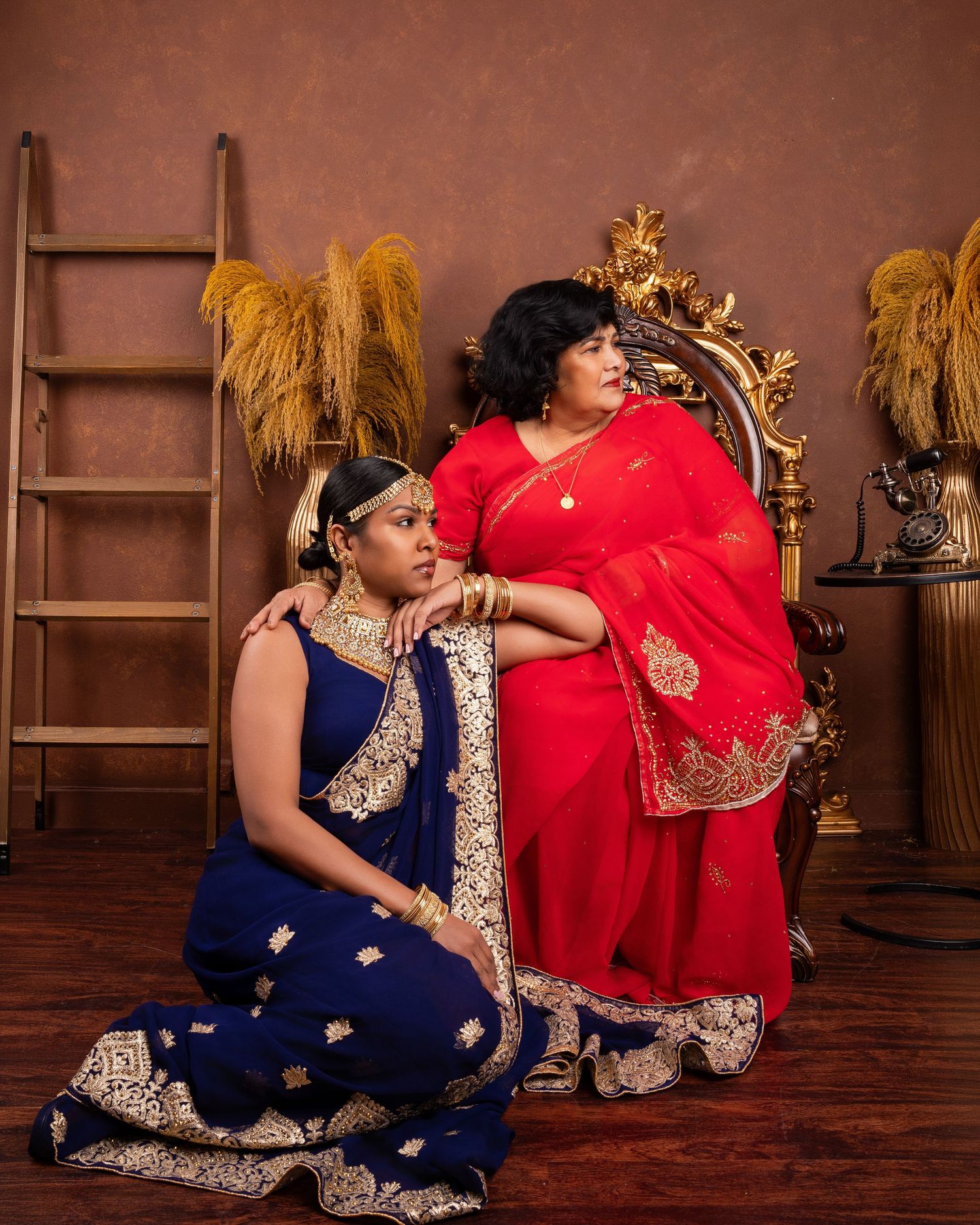 A person in a navy blue and gold sari poses gracefully on a brown studio floor with decorative wheat stalks in the background.