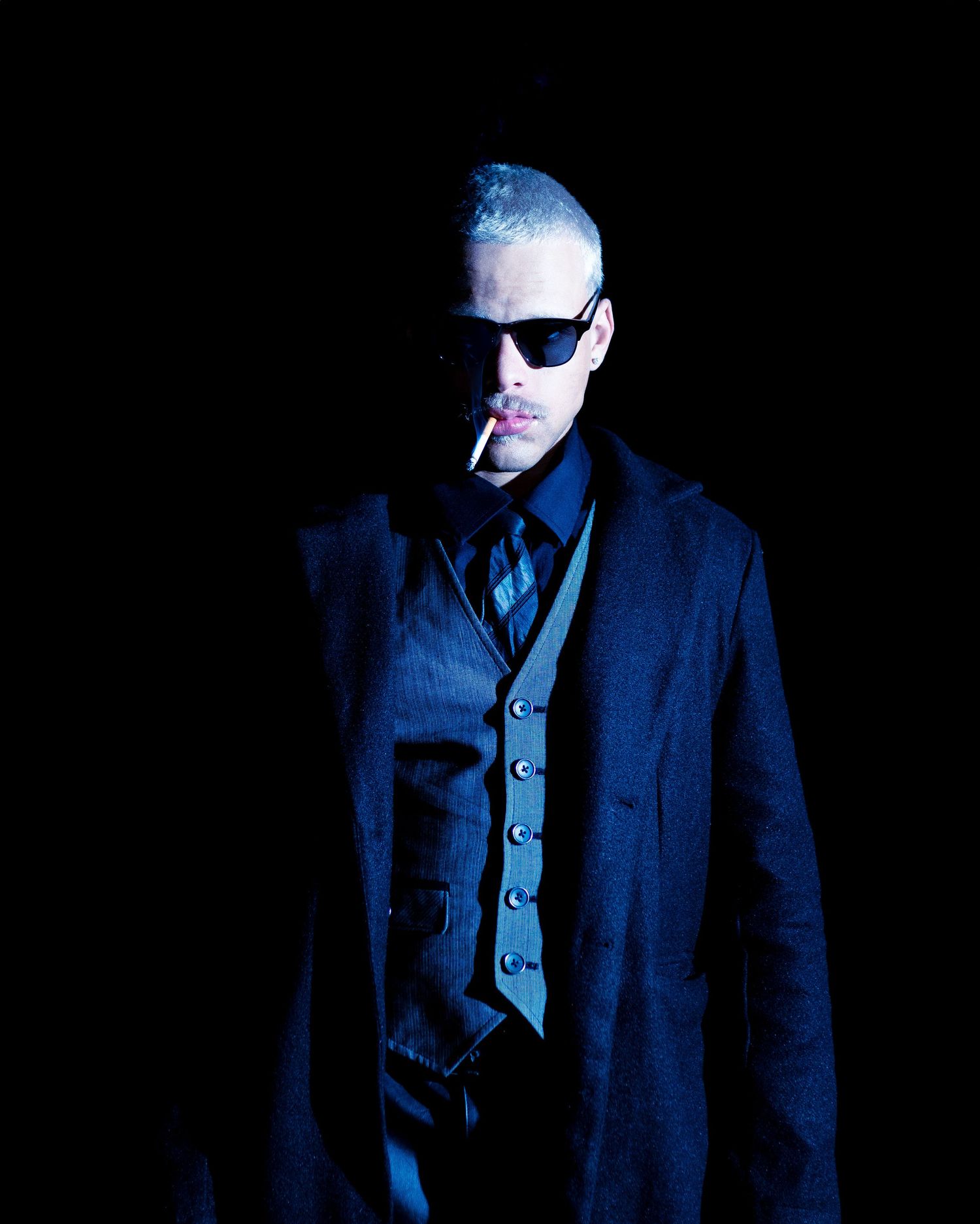 A person in sunglasses and navy suit poses dramatically against dark background with moody lighting.