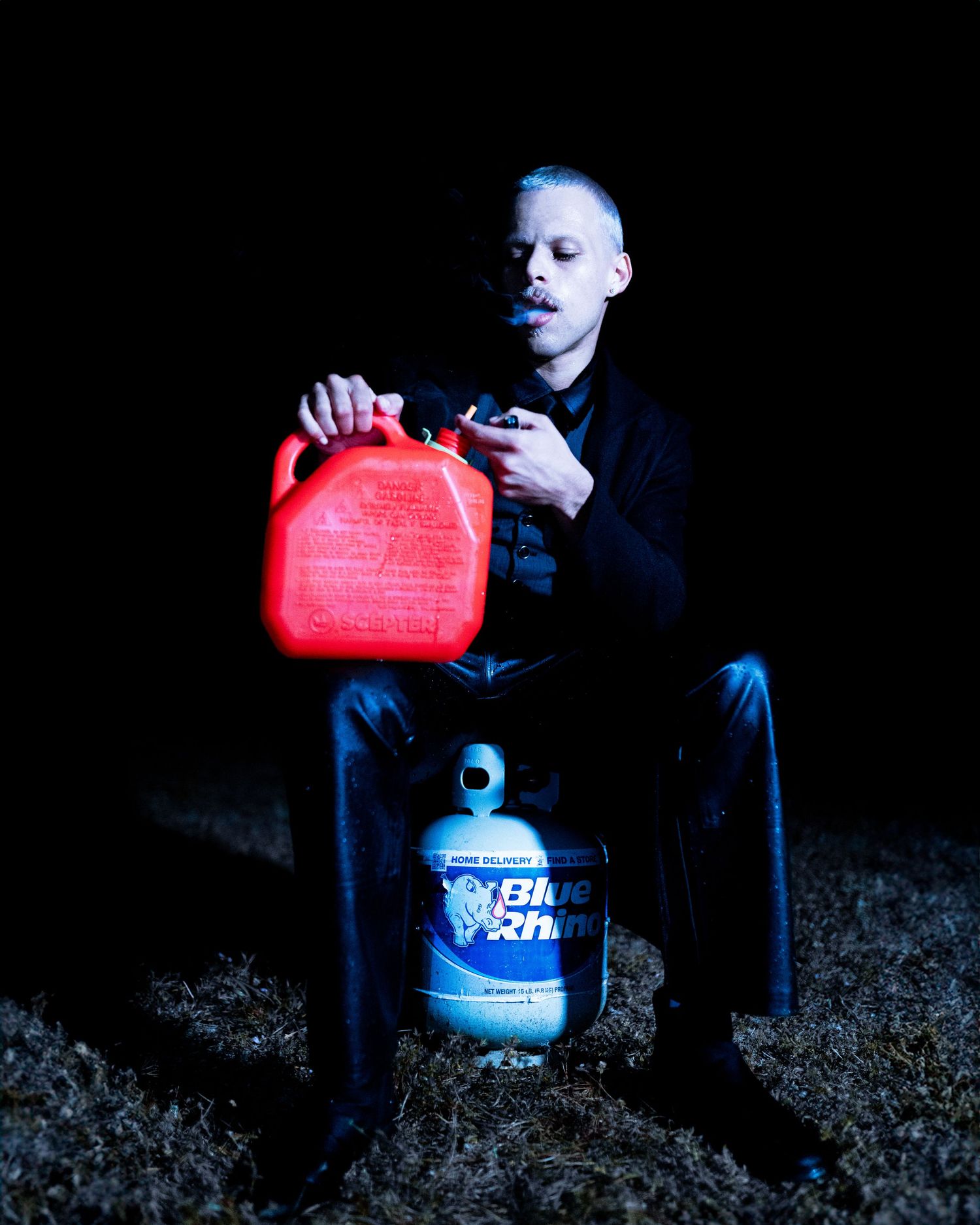 A figure in dark clothing sits with red gas can and blue container against black background.