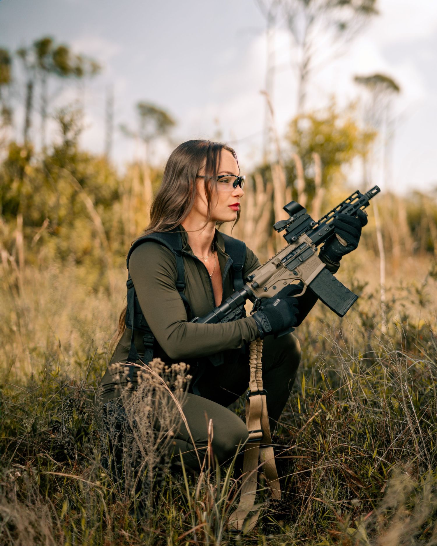 A person in tactical gear holds a military-style rifle while crouching in tall grass during an outdoor photoshoot.
