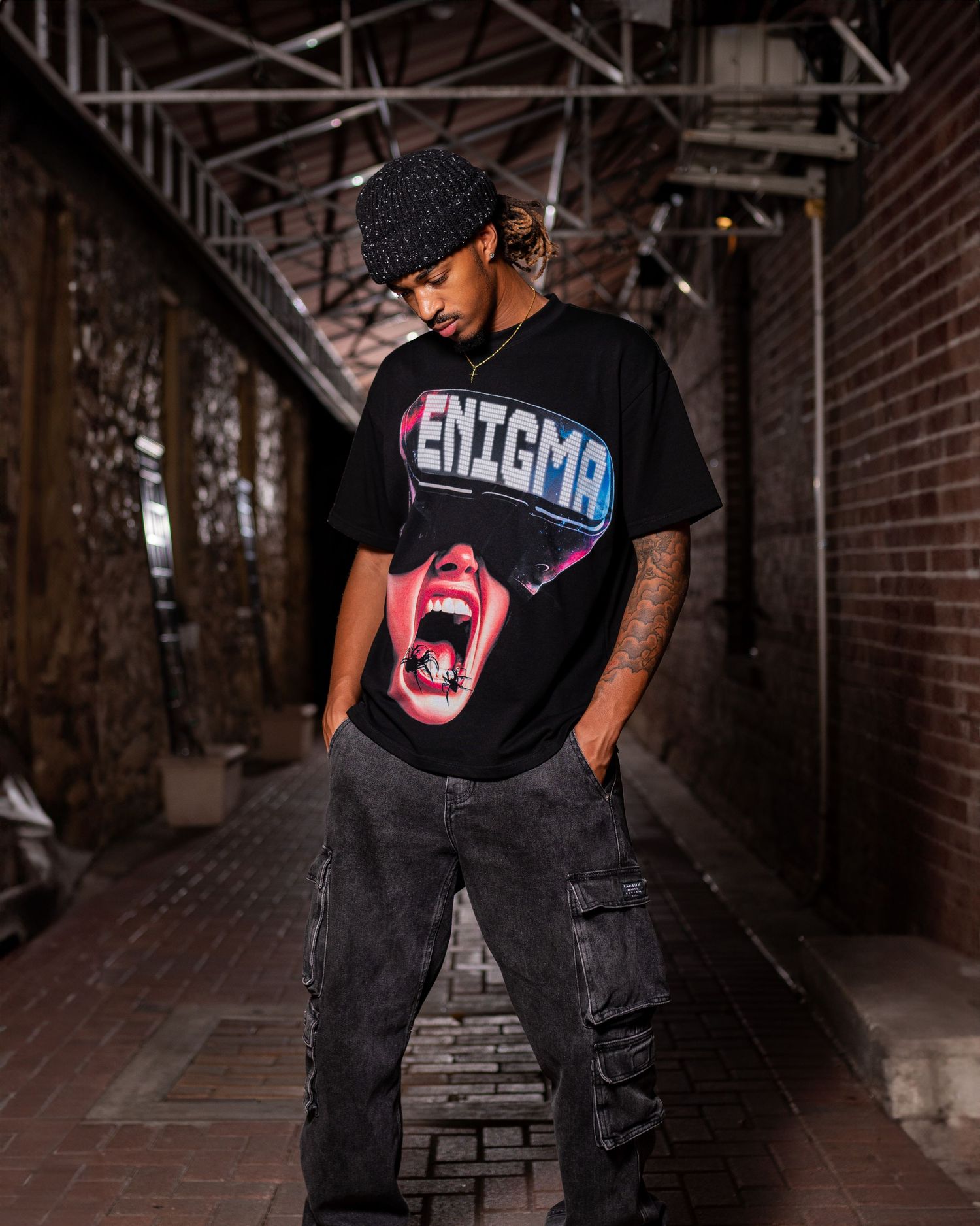 Person wearing black Enigma t-shirt with screaming mouth graphic poses in urban alley setting.