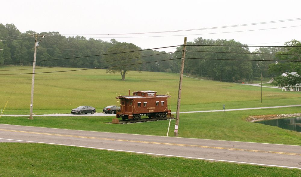 A red caboose sits on railroad tracks in a green rural landscape with trees in the background.